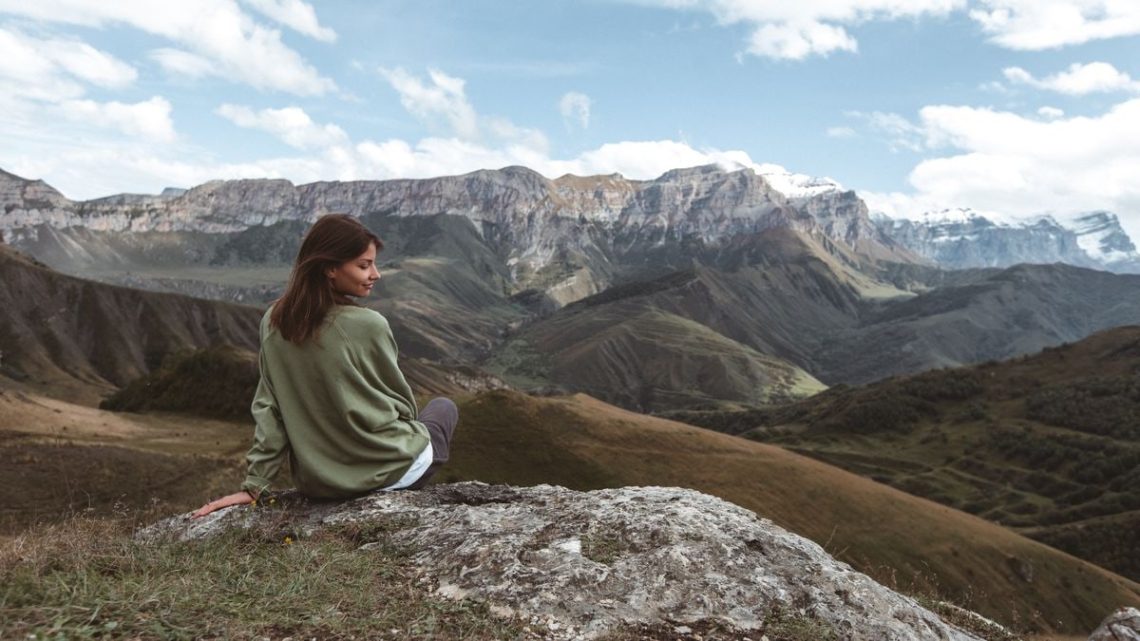 A woman in a green sweater sits on a rock, enjoying the stunning view of majestic mountains during the day.