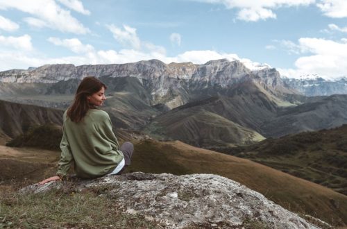 A woman in a green sweater sits on a rock, enjoying the stunning view of majestic mountains during the day.